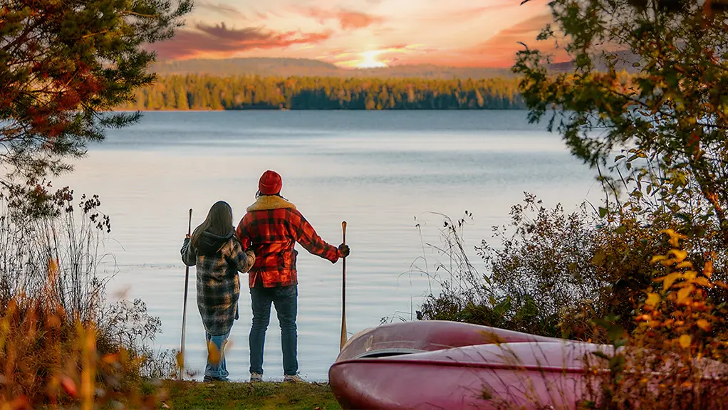 Dos personas de pie a la orilla de un lago tranquilo, con una canoa y un remo, rodeadas de los colores del otoño.