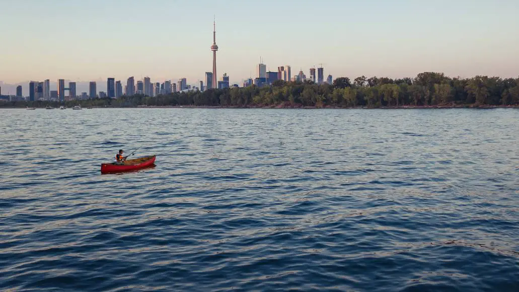 A man cast a fishing line from a canoe in the waters of Toronto’s harbour, with the CN Tower and city skyline in the background.