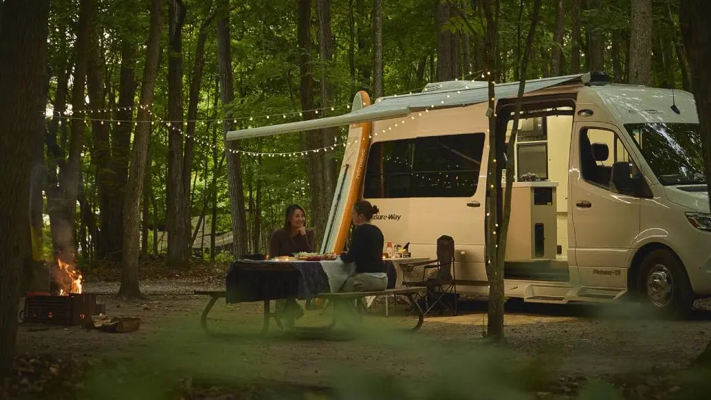 Zwei Frauen genießen ein Abendessen an einem Picknicktisch neben einem Wohnmobil, das auf einem Campingplatz abgestellt ist.