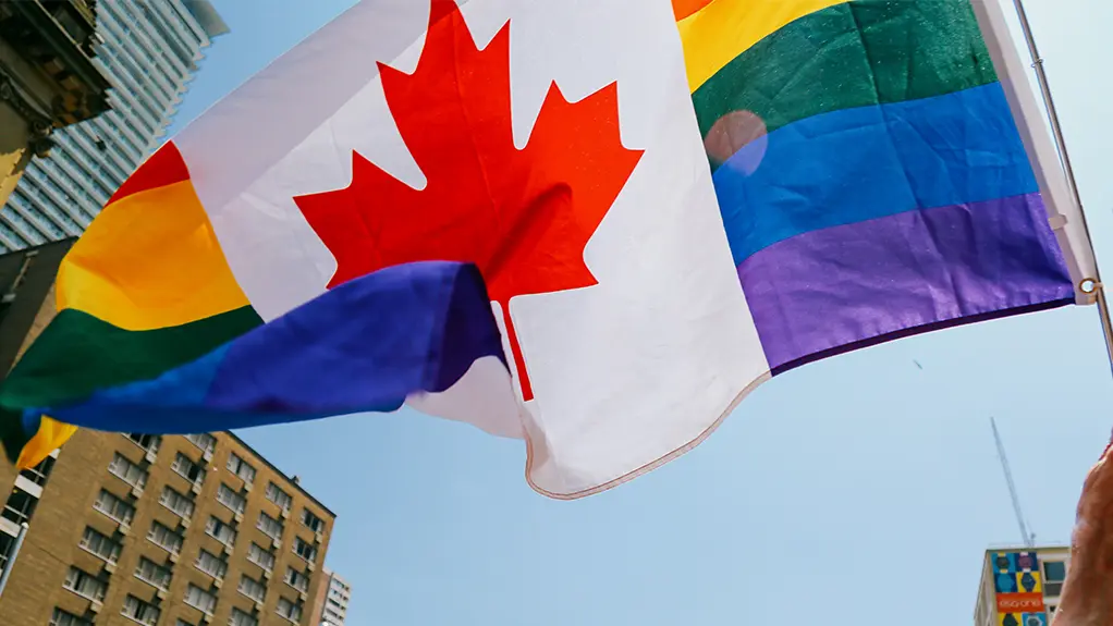 Menschen schwenken Regenbogen- und kanadische Flaggen während der Toronto Pride Parade auf einer Straße der Stadt, umgeben von farbenfrohen Festivalbesuchern und im Sonnenlicht.