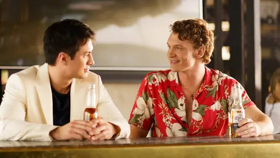Deux hommes assis à un bar tenant des bouteilles de bière, discutant et se souriant l’un à l’autre, l’un portant un blazer blanc et l’autre, une chemise rouge à fleurs, dans un cadre moderne et chaleureux.