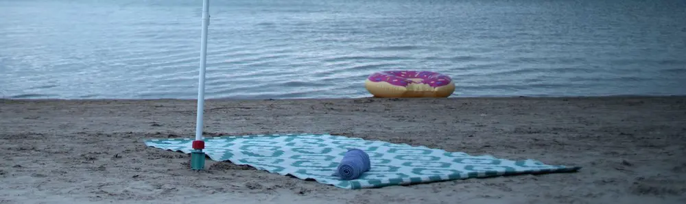 Ein bunter Sonnenschirm spendet Schatten über einem Handtuch an einem Sandstrand am Seeufer.