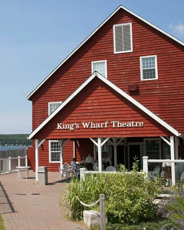 People enjoying a conversation outside of a red, wooden building that is located beside the waterfront