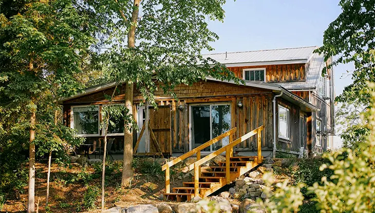 Cabaña rústica de madera rodeada de árboles con una pequeña escalera que conduce a la entrada.